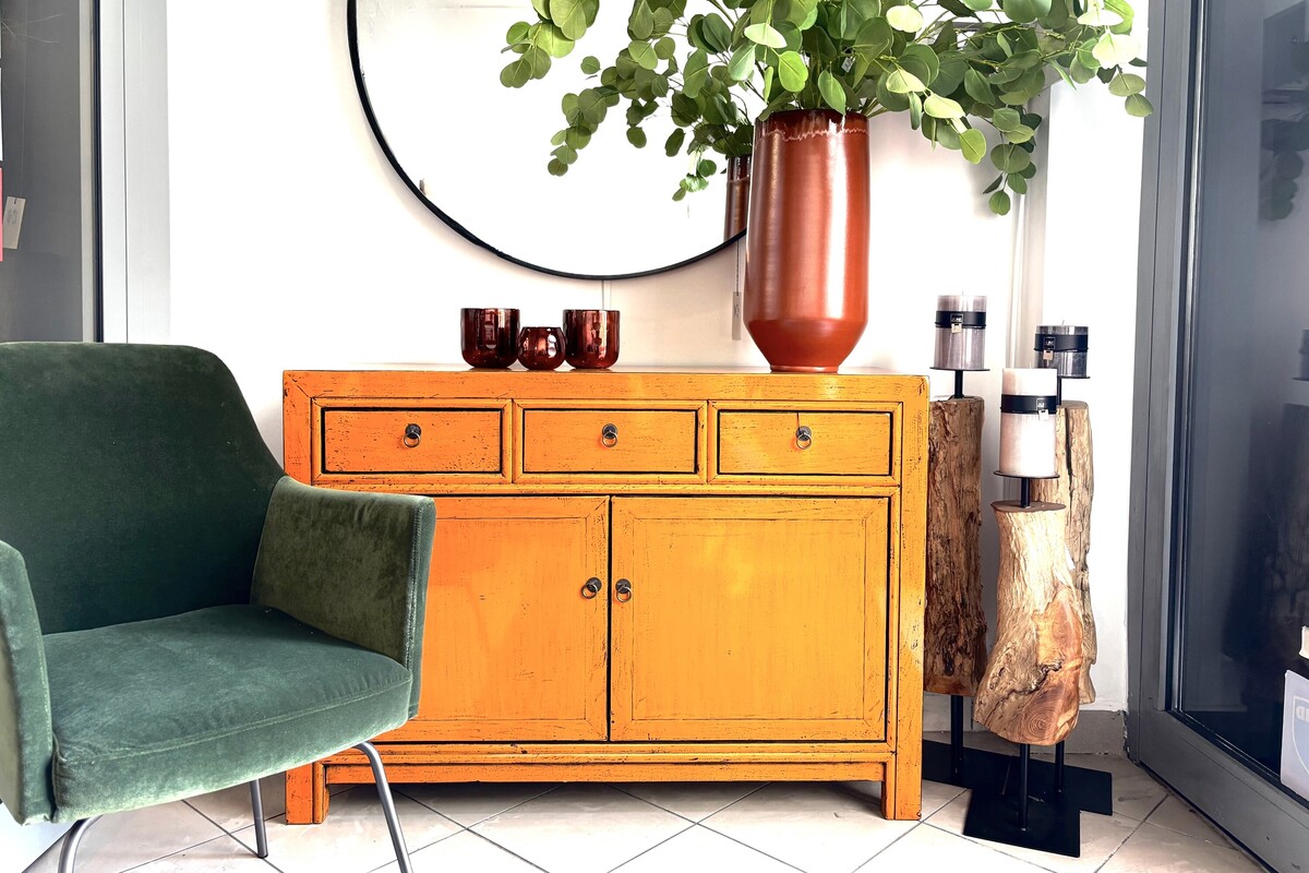 Pomarańczowa komoda. Biała ściana. Aranżacja hollu ciekawe dekoracje drewniane. Ceramiczny wazon, sztuczne liście eucaliptusa.