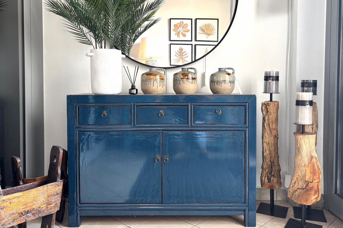 Granatowa komoda na białej ścianie. Aranżacja hollu. Okrągłe lustro w czarnej metalowej ramie. Drewniane świeczniki. 
